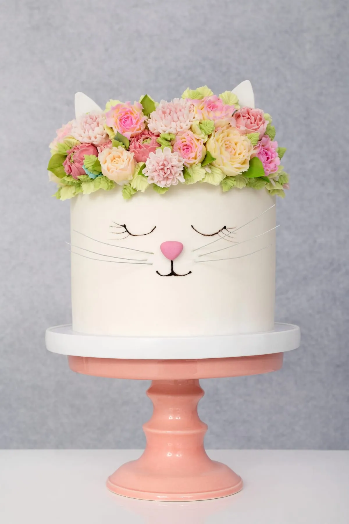 A white cat cake with a crown of buttercream flowers and pink ears